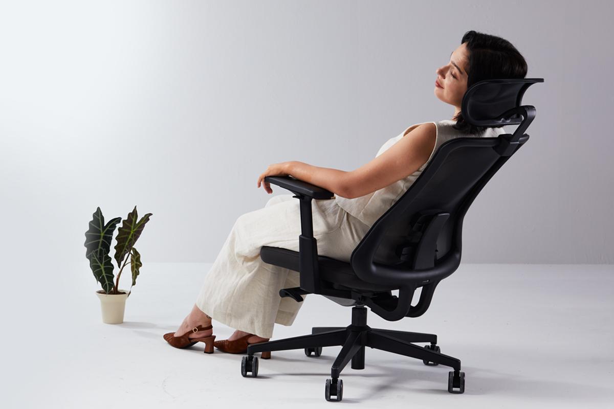 Woman in white outfit reclining in black ergonomic office chair demonstrating full recline position with headrest support and proper spine alignment on white backgroun