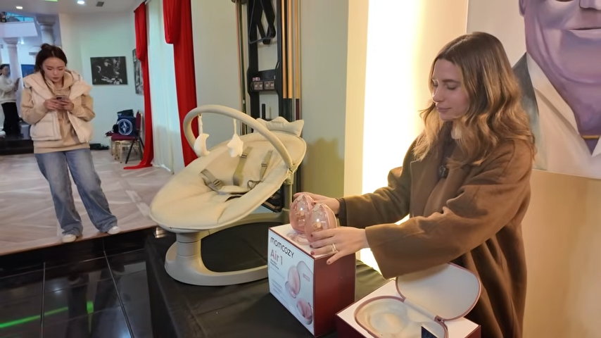 Nicolas Charbonnier in a brown coat unboxing and displaying the rose gold Momcozy Air 1 wearable breast pump next to a beige baby swing at an event